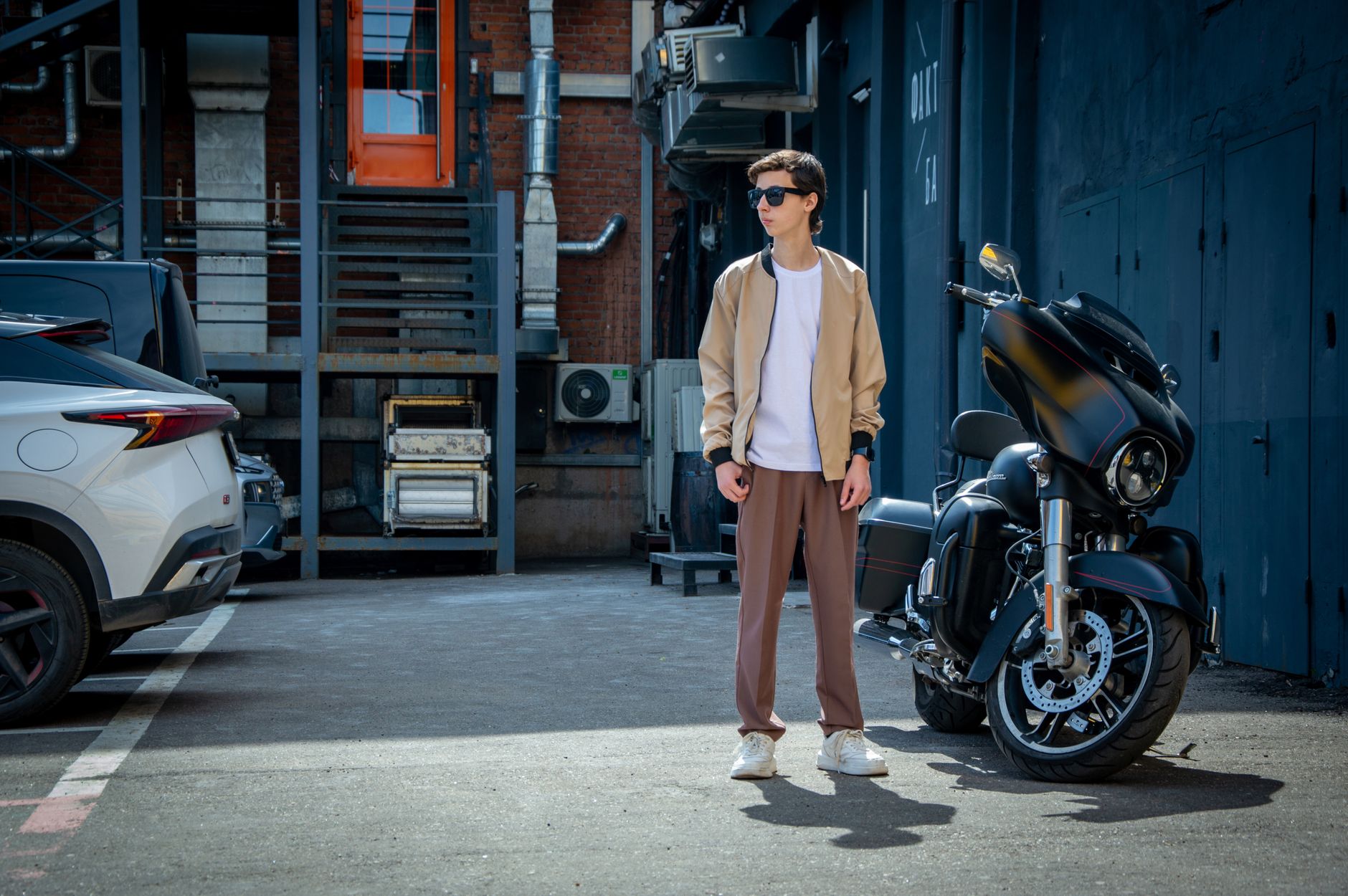 Jeune homme en pantalon chino marron et bombers beige dans une ruelle urbaine, à côté d'une moto.